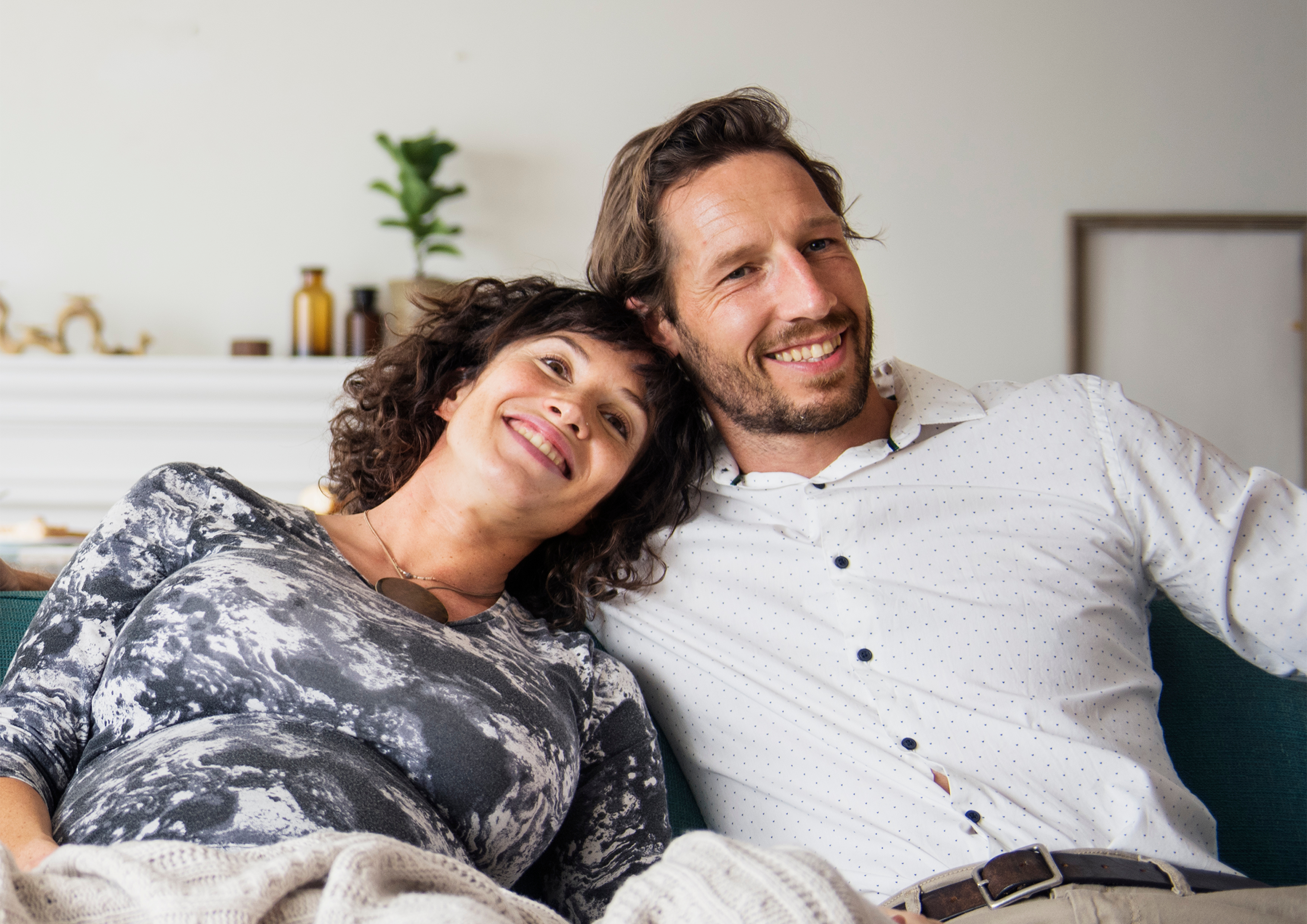 Smiling woman and man sitting closely on couch, woman goes to gala with client as his date, husband surprised emotions
