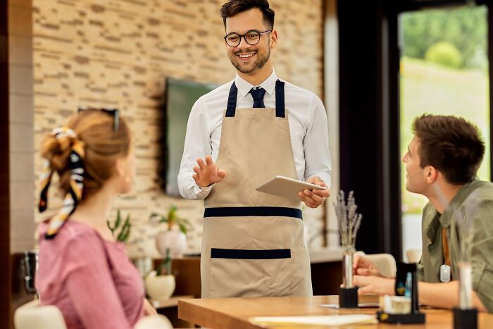 Young waiter with apron and tablet smiling while talking to a confused couple, showing moments of misreading social cues.