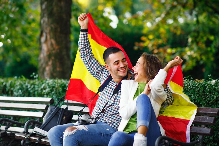 Couple smiling on park bench with Spanish flag, sharing unbelievable facts about their country that are actually true.