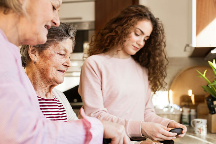 Three women in a kitchen, two older and one younger, sharing moments and reflecting on life’s hardest truths together.