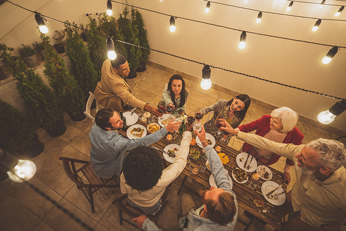 Group of friends enjoying dinner party outdoors with string lights, raising glasses in a toast, capturing obnoxious dinner guest husband dread.