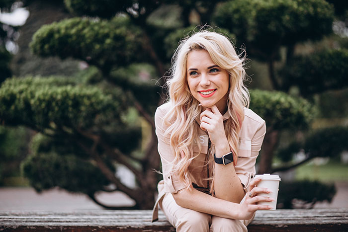 Blonde woman smiling outdoors sitting on bench with coffee cup, representing famous supermodel lifestyle and success.
