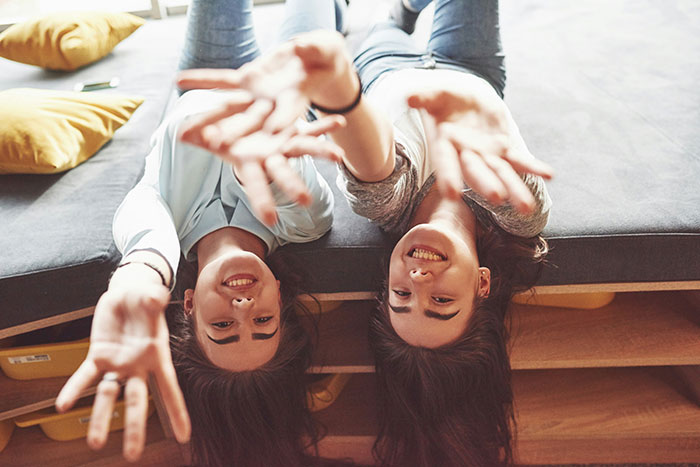 Two smiling young women lying upside down on a couch reaching out, representing famous supermodels in a casual setting.