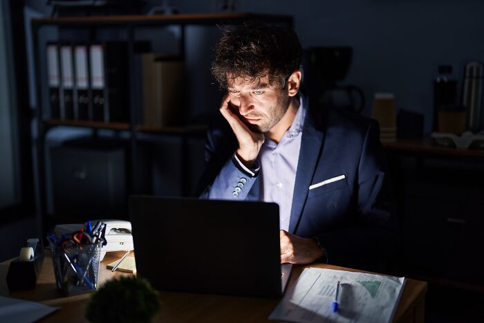 Accountant working late at desk, focused on laptop, surrounded by office supplies and paperwork in dimly lit room.