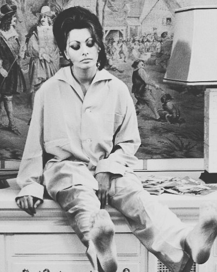 Sophia Loren seated barefoot in relaxed attire in front of a detailed mural, showcasing Italian beauty and Hollywood legend.