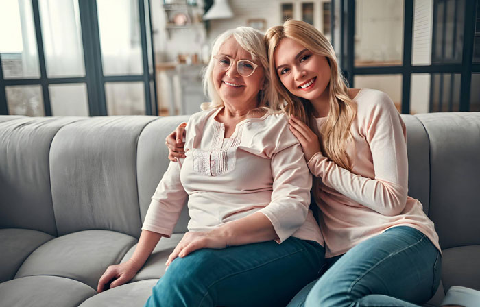 Pregnant woman sitting with her supportive mother at home, smiling and embracing on a gray sofa in a bright living room. Pregnant woman sitting with her supportive mother at home, smiling and embracing on a gray sofa in a bright living room.