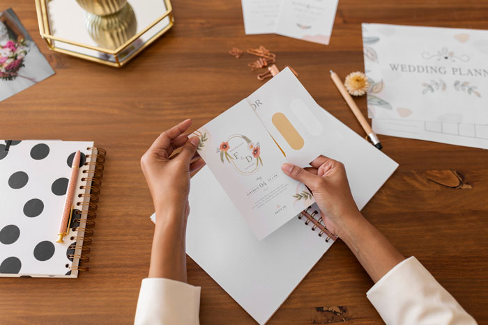 Person holding a wedding invitation card at a wooden table with wedding planning materials and notebooks nearby.