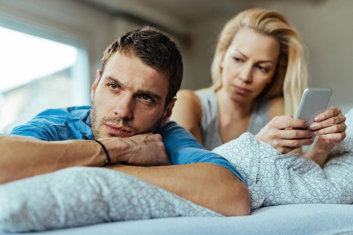 Couple on bed looking upset while woman holds phone, illustrating selfish SIL trying to ruin couple&rsquo;s honeymoon conflict.