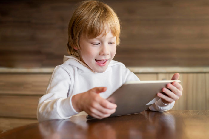 Young boy sitting at a wooden table, focused on a tablet, illustrating hurdles to eating together with family.