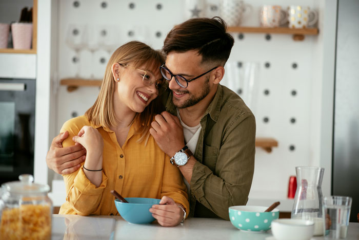 Young couple smiling and embracing in kitchen as mom struggles with challenges to eat together with family.