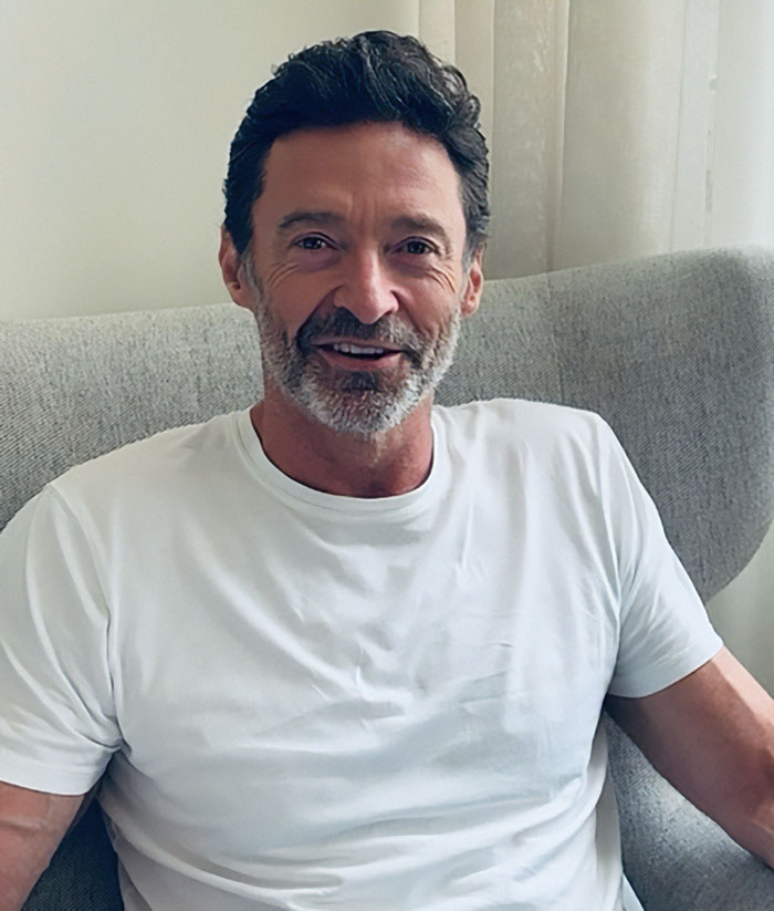 Hugh Jackman sitting on a gray chair in a white shirt with a relaxed expression during midlife crisis period.