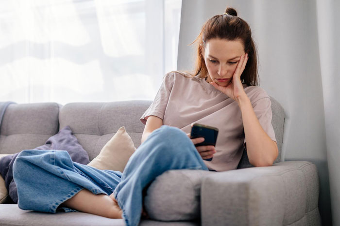 Young woman looking worried on couch, holding phone, reflecting concern about slacker hubby plans after wife's surgery.