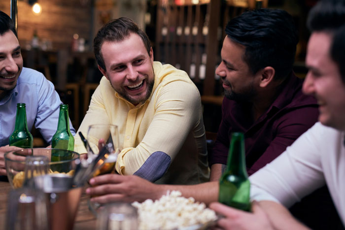 Group of men casually chatting and drinking at a bar, reflecting slacker hubby planning to skip caring for wife after surgery.