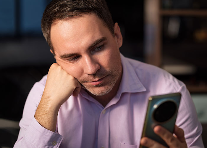 Man in a pink shirt looking thoughtfully at his smartphone, reflecting on a woman&rsquo;s dream of having a tummy tuck.
