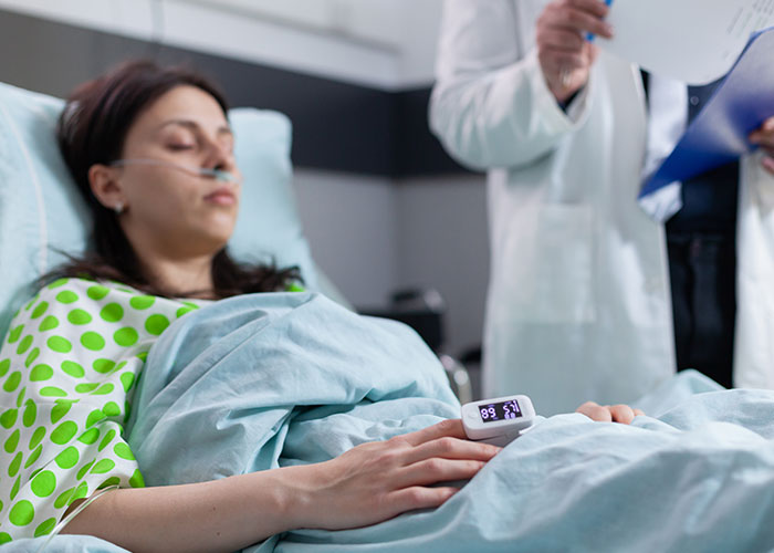 Woman lying in hospital bed with oxygen tube and fingertip pulse oximeter, relating to tummy tuck savings loss story