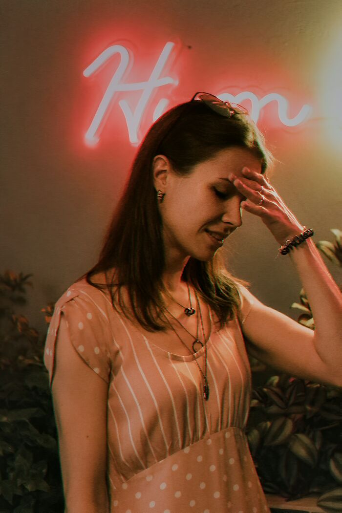 Young woman in a soft pink dress showing embarrassment by holding her forehead in a dimly lit setting with neon lights.