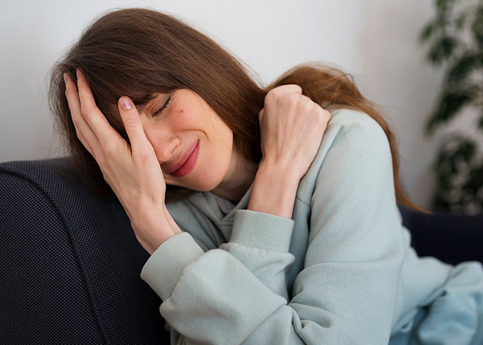 Young woman in a light blue sweatshirt sitting on a couch, showing a shy smile and covering part of her face with her hand.