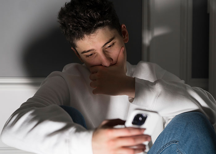 Young man in white sweatshirt looking upset while holding a phone, reflecting on icks that make men lose interest in a woman.