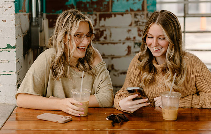 Two women smiling and enjoying drinks while looking at a phone, reflecting woman revenge on cheating boyfriend online.