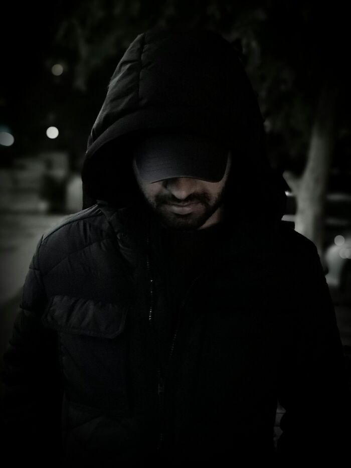 Man in dark hooded jacket and cap standing outside at night, evoking a true crime fan atmosphere.
