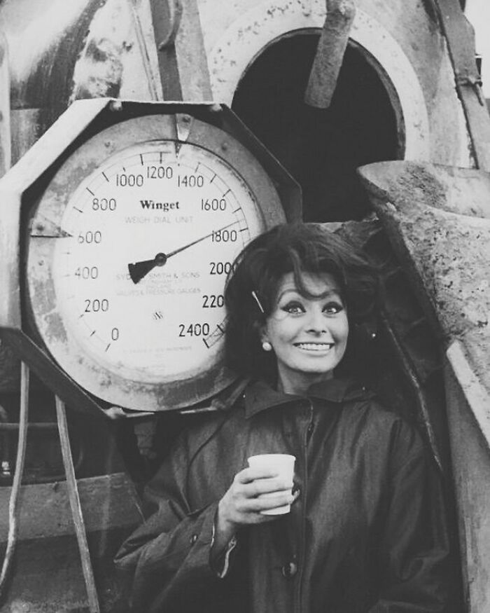 Sophia Loren smiling and holding a cup near a large vintage weighing scale, showcasing Italian beauty and Hollywood charm.