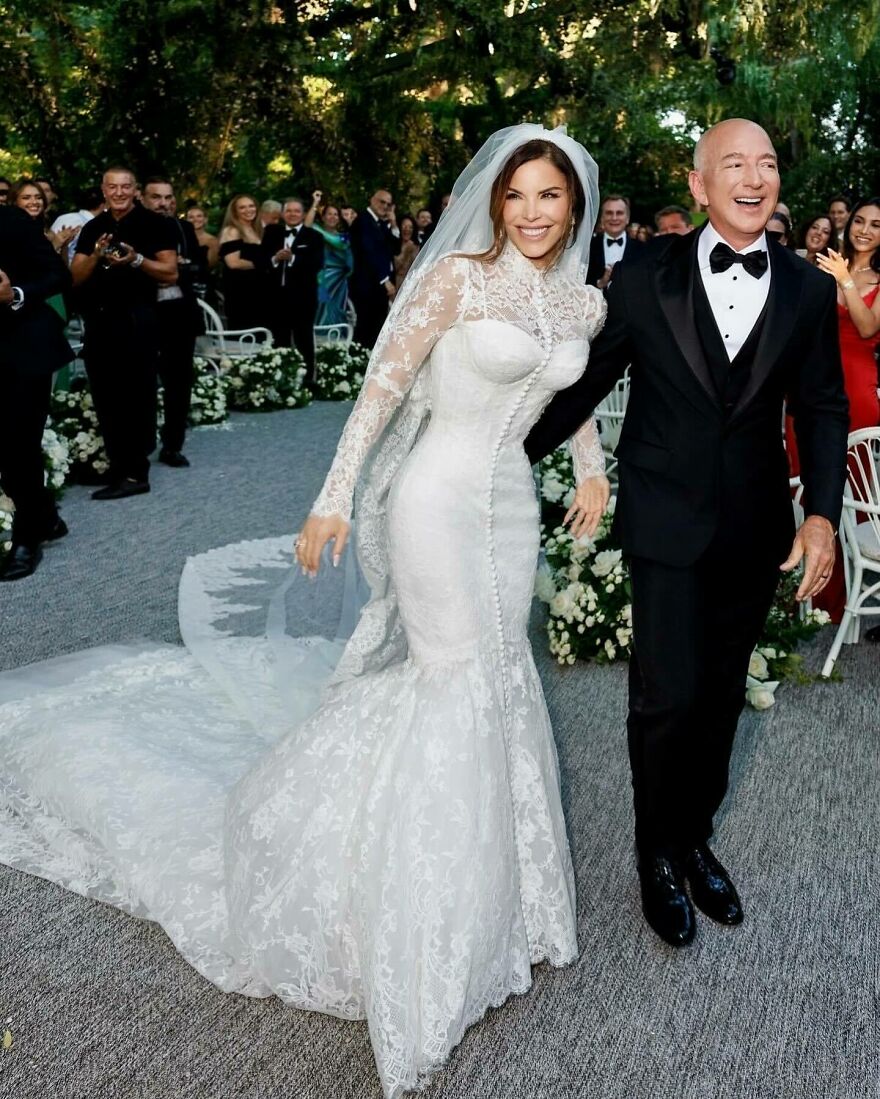 Lauren S&aacute;nchez in a lace wedding gown smiling next to a man in a black tuxedo during an outdoor ceremony.