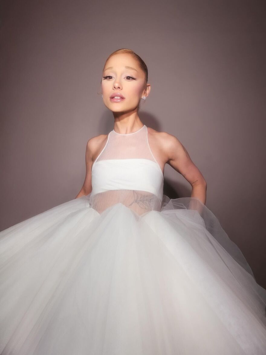 Ariana Grande posing in a white sheer gown with a voluminous skirt, showcasing a soft makeup look and sleek hairstyle.