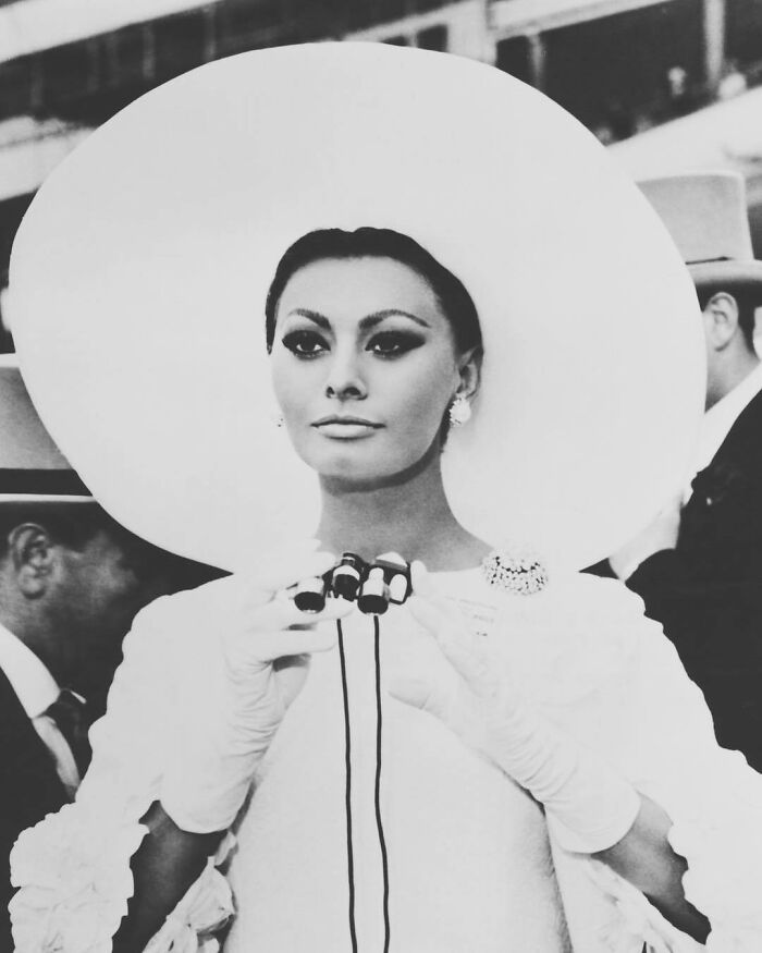 Sophia Loren wearing a large hat and gloves, holding opera glasses, showcasing Italian beauty and Hollywood legend style.
