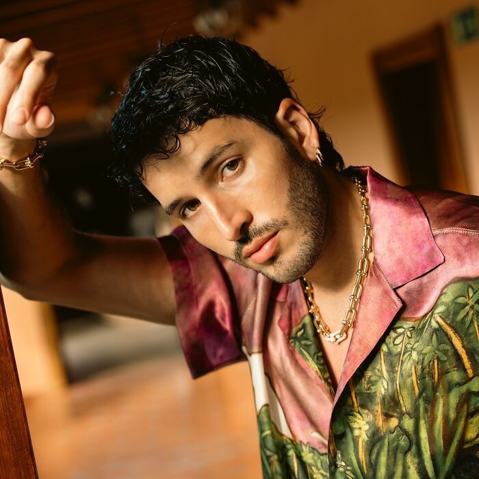 Man with curly hair and gold chain wearing a tropical shirt posing indoors for most handsome man in the world.