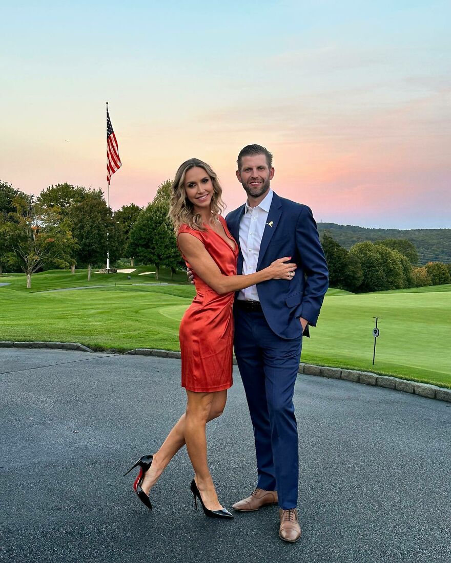 Lara Trump in a red dress with a man in a navy suit posing outdoors at sunset on a golf course background.