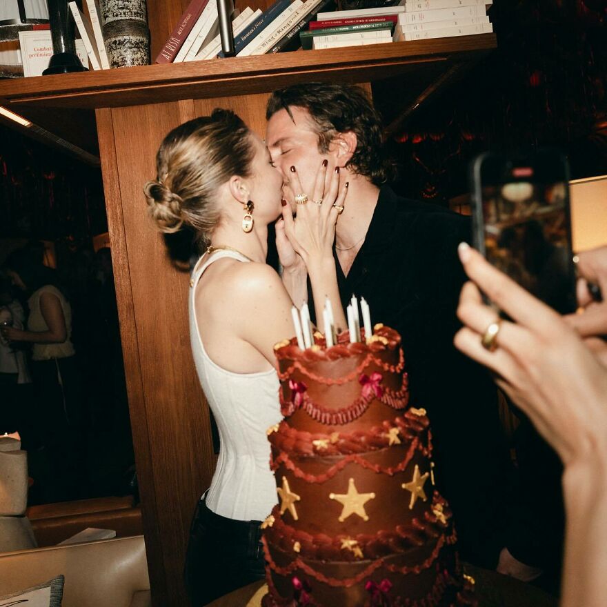 Gigi Hadid and Bradley Cooper sharing a kiss at a celebration with a decorated cake and guests capturing the moment.