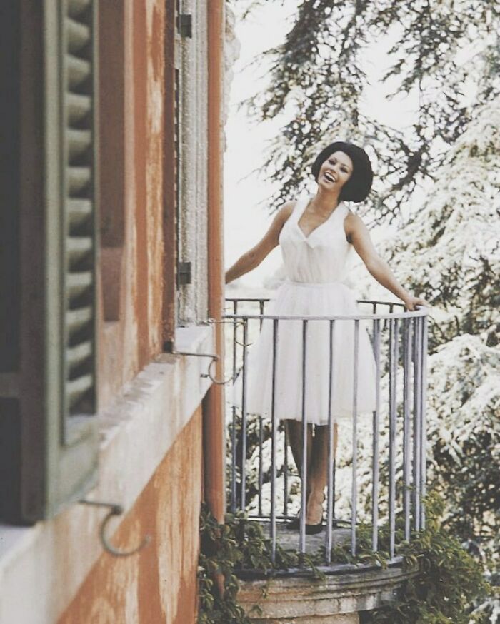 Sophia Loren smiling joyfully on a small balcony wearing a white dress, capturing Italian beauty and Hollywood legend charm.