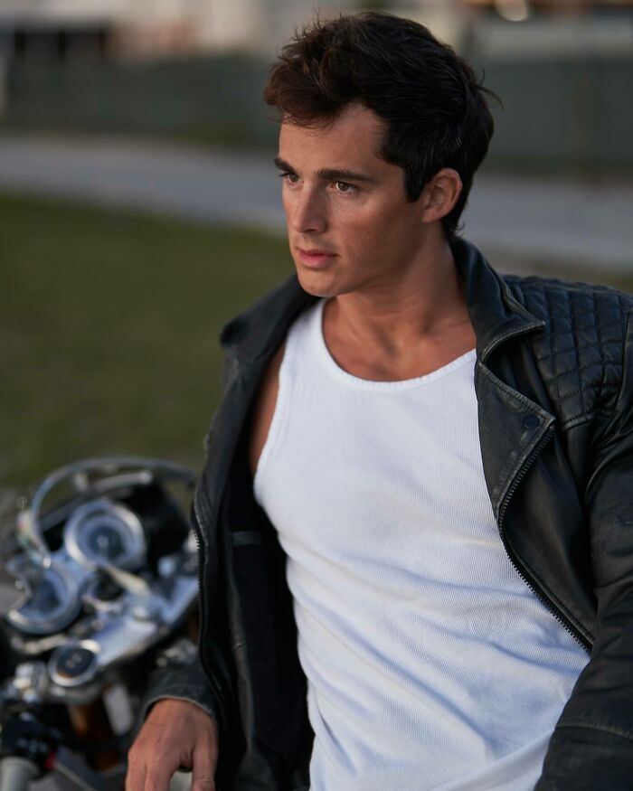 Young man wearing a leather jacket and white tank top, posing outdoors by a motorcycle, embodying handsome man style.