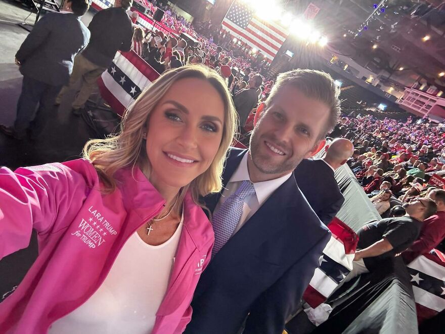 Lara Trump smiling at a political rally wearing pink jacket with crowd and American flags in the background.