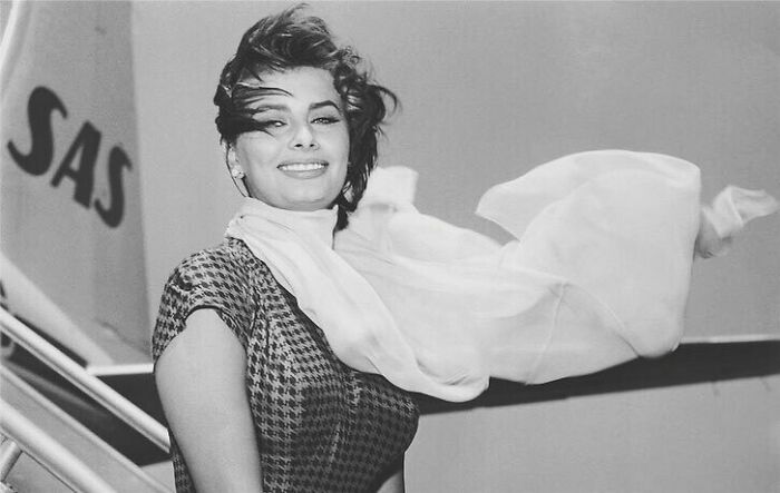 Sophia Loren smiling with a flowing scarf, standing near an airplane, showcasing Italian beauty and classic Hollywood charm.