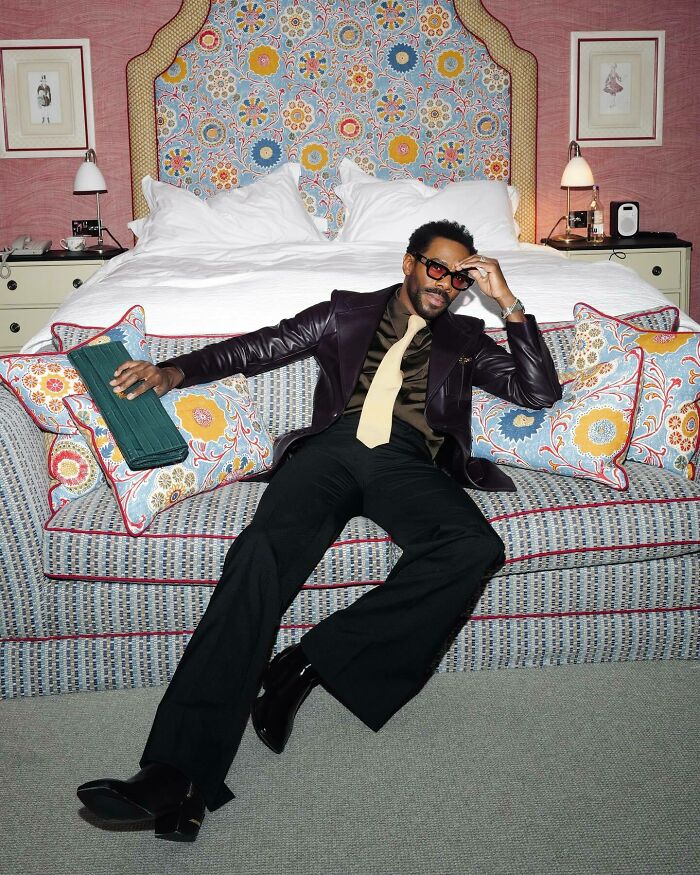 Stylish man posing on a patterned couch in a suit and sunglasses, embodying the most handsome man vibe 2025.