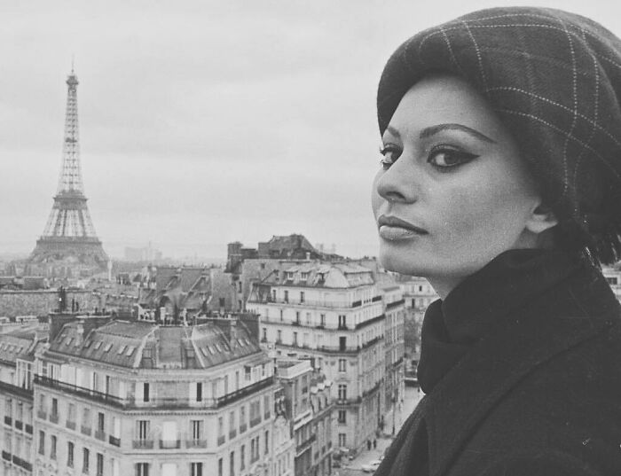 Sophia Loren in Paris wearing a beret, with the Eiffel Tower and classic buildings in the background, showcasing Italian beauty.