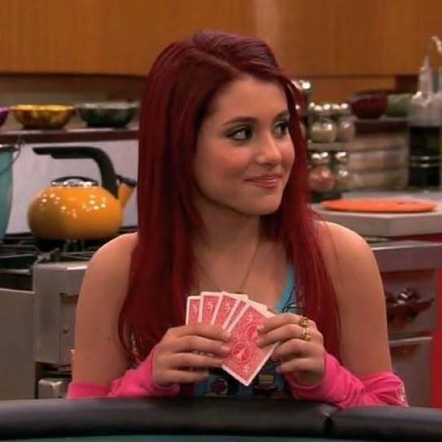 Ariana Grande with red hair holding playing cards, smiling in a kitchen setting during an early career phase.