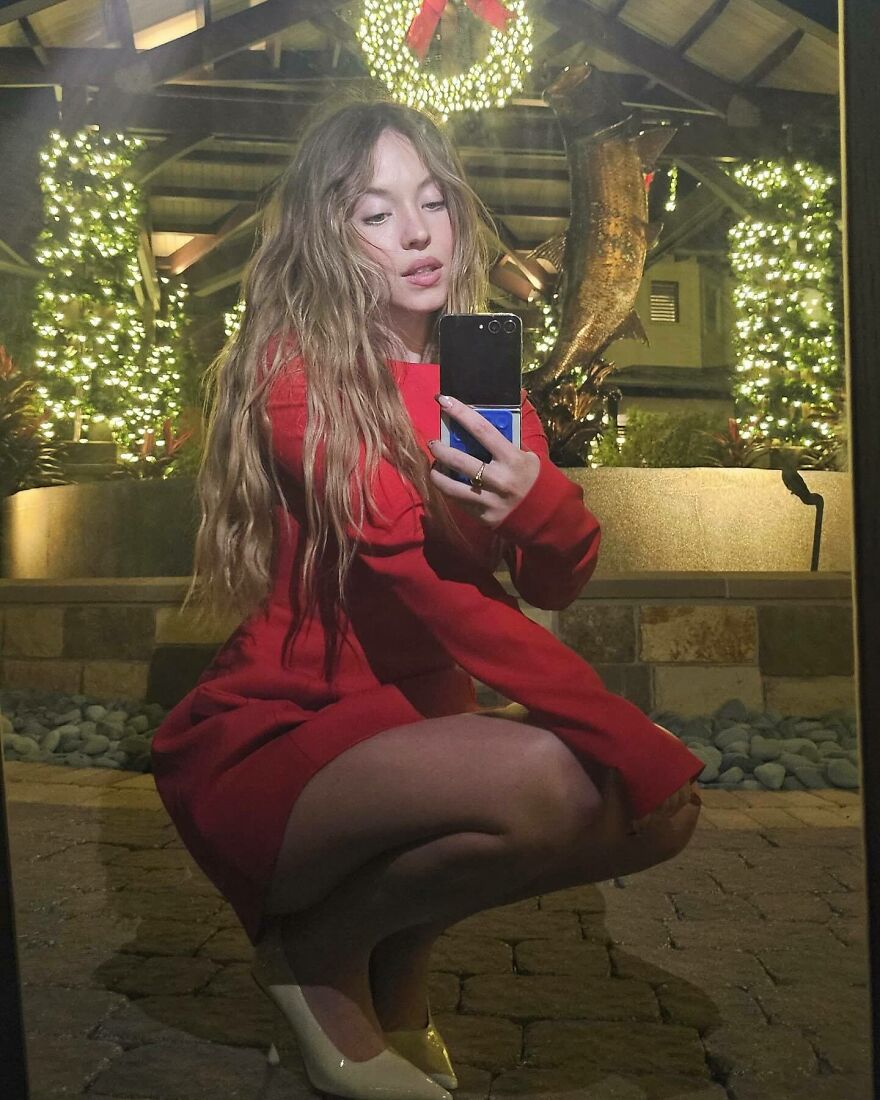 Sydney Sweeney taking a mirror selfie in a red dress at night with festive lights illuminating the background. Sydney Sweeney taking a mirror selfie in a red dress at night with festive lights illuminating the background.