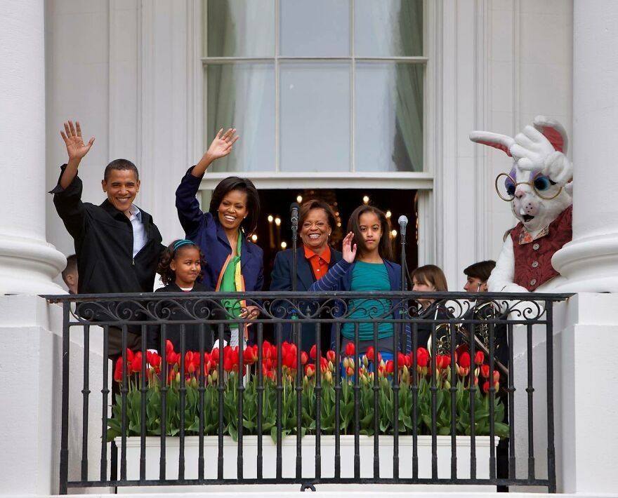 Michelle Obama smiling and waving on a balcony with family members and a person in a bunny costume behind flowers. Michelle Obama smiling and waving on a balcony with family members and a person in a bunny costume behind flowers.