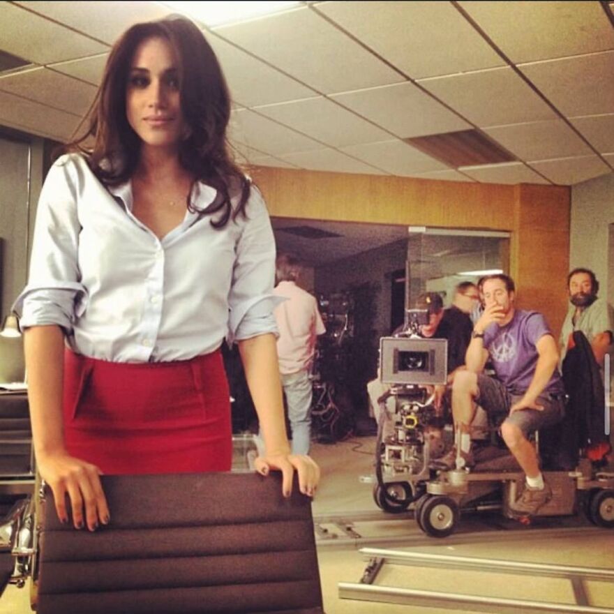 Meghan Markle on a film set wearing a white shirt and red skirt with crew and camera equipment in the background. Meghan Markle on a film set wearing a white shirt and red skirt with crew and camera equipment in the background.