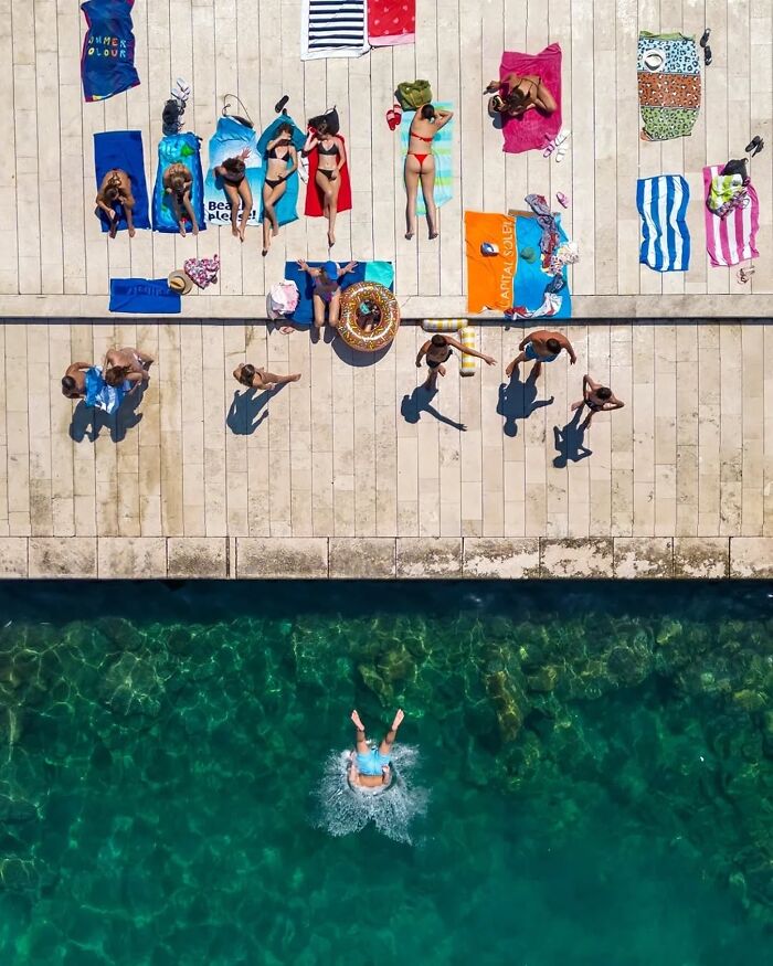 Aerial view of people sunbathing on colorful towels and one person diving into clear water, showcasing fascinating photos.