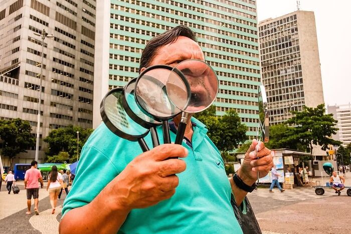 Man holding multiple magnifying glasses distorting his face in an urban setting with tall buildings and pedestrians nearby.