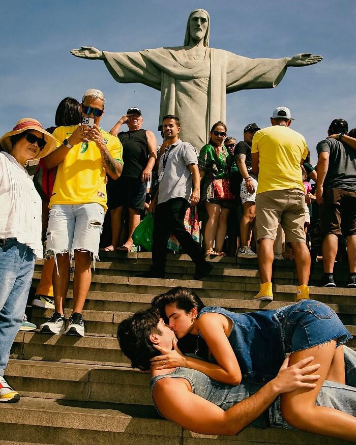 Couple kissing on stairs in front of Christ the Redeemer statue with a crowd, a fascinating photo capturing a unique moment.