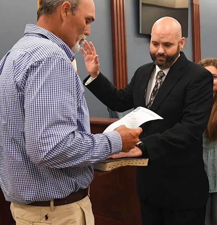 Police chief in black suit raising hand for oath while another man in checkered shirt reads a document indoors.