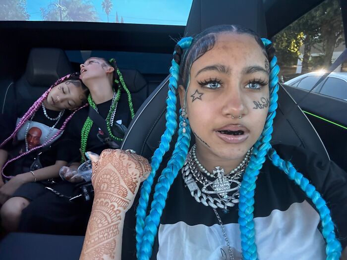 Young woman with blue braids, fake face tattoos, piercings, and grills sitting in a car with two others asleep in the back.