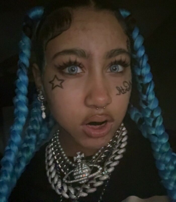 North West with fake face tattoos, piercings, grills, and blue braids in a dimly lit setting.
