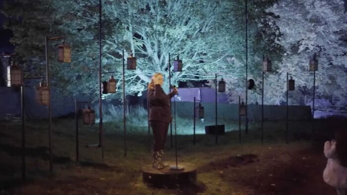 Person standing on a round platform reading from a microphone surrounded by lanterns in a nighttime outdoor setting.