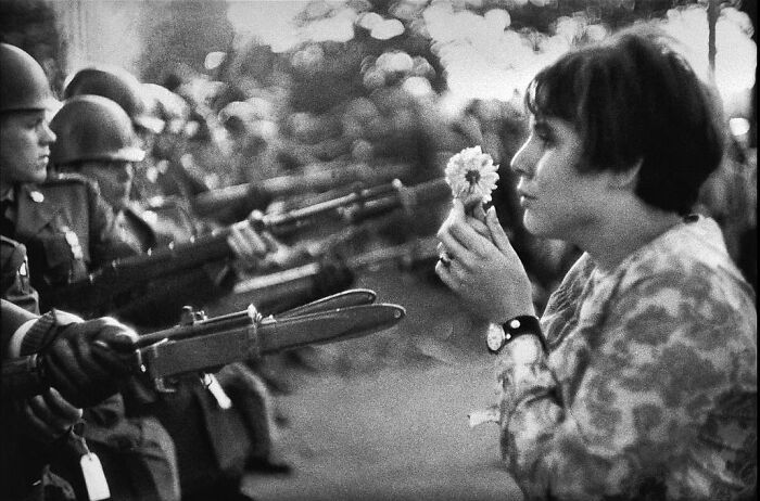 Black and white photo showing a peaceful woman holding a flower facing soldiers with bayonets, a fascinating historical moment captured.