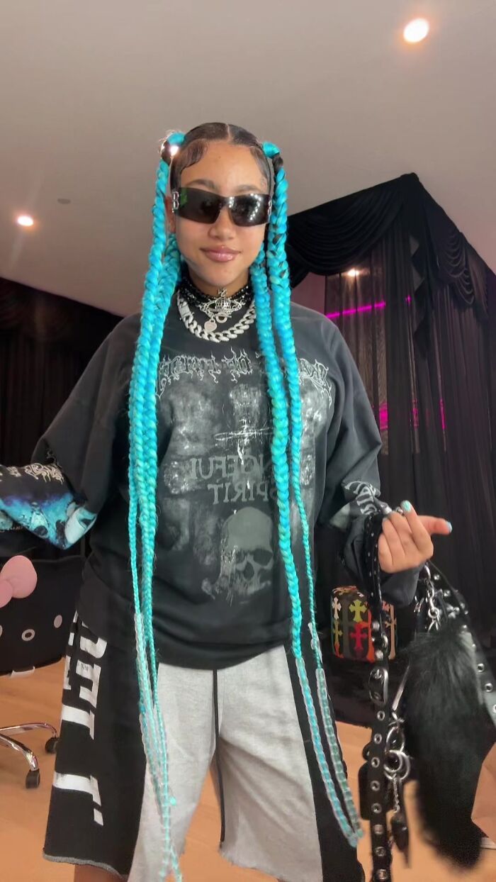 Young woman with long blue braids wearing sunglasses, chains, and holding accessories, showing fake face tattoos and piercings.